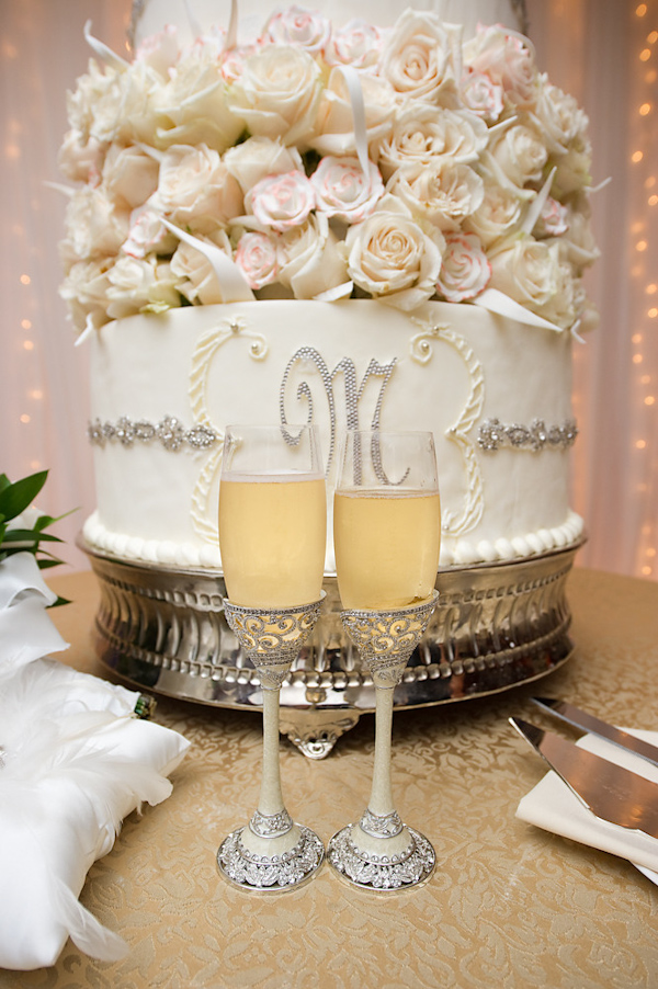 gorgeous champagne round wedding cake with rhinestone border and rhinestone monogram design and ivory and light pink roses on top - cake is on a vintage silver cakestand sitting on table with a gold tablecloth and two vintage style ivory champagne flutes with rhinestone design  -  photo by Houston based wedding photographer Adam Nyholt